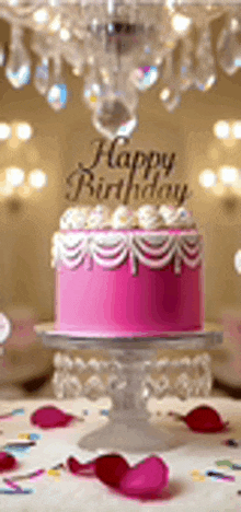 a pink birthday cake is on a cake stand on a table with petals .