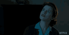 a woman in a blue shirt and pearls looks up at something in the dark