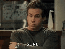 a man in a gray shirt is sitting at a table with his arms crossed and says `` sure '' .