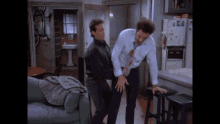 two men are standing next to each other in a living room with a refrigerator in the background .