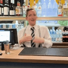 a bartender wearing suspenders and a tie stands behind a counter with bottles of bacardi and smirnoff on it