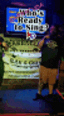 a man is singing into a microphone in front of a sign that says who 's ready to sing ?