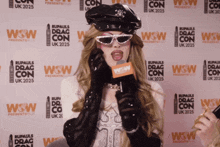 a woman speaking into a microphone in front of a wall that says rupauls drag con