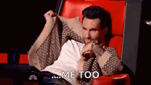 a man is sitting in a red chair with his sweater over his shoulder and says `` me too '' .