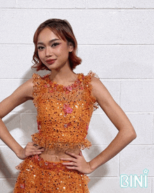 a woman in an orange dress is standing in front of a white brick wall and the word bini is on the bottom