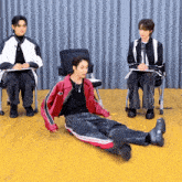 a man in a red jacket is laying on the floor while two other men sit in chairs behind him .