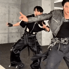 two men are standing next to each other in a room . one of the men is wearing a leather jacket .
