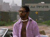 a man in a purple shirt and sunglasses is standing in front of a car .