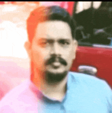 a man with a beard and mustache is wearing a blue shirt and standing in front of a red car .