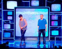 two men are standing on a stage in front of a wall of televisions
