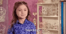 a little girl in a blue shirt is standing in front of a shelf with snickers on it .