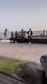 a group of people are standing on a brick walkway near a body of water with a viggle.ai logo in the corner