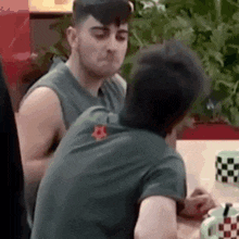 two men are sitting at a table talking to each other . one of the men has a star on his shirt .