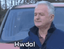 a man in a blue jacket is standing in front of a van and says hwda !