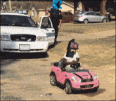 a girl is driving a pink toy car with a police officer standing behind her