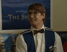 a man wearing glasses and a bow tie is smiling in front of a poster that says the big