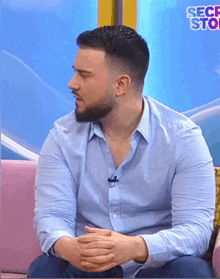 a man in a blue shirt sits on a pink couch with his hands folded