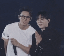 two men are standing next to each other on a stage . one of the men is wearing glasses and holding a microphone .