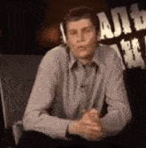 a man in a grey shirt is sitting in a chair with his hands folded and talking .
