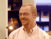 a man wearing glasses is smiling in front of a bookshelf with a book called heal on it .