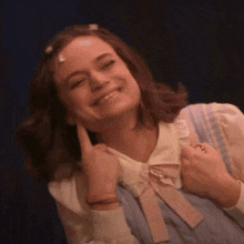 a woman is smiling and touching her ear while wearing a pink dress and a white shirt .
