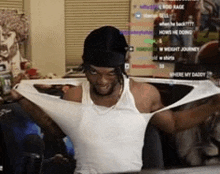 a man is wearing a white tank top and a black hat while sitting in front of a computer screen .