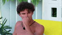 a shirtless man with curly hair is sitting on a yellow couch and covering his nose with his hand .