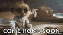 a meerkat doll is sitting on a table and says `` come home soon '' .