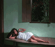 a woman in a white dress is laying on a wooden bench in front of a window