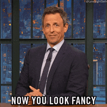 a man in a suit and tie is sitting at a table and says now you look fancy