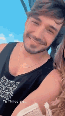 a man with a beard wearing a black tank top is smiling while standing next to a woman with long hair .