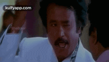 a man with a mustache is wearing a white suit and tie and making a funny face .