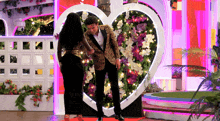 a man and a woman are standing in front of a heart shaped wall of flowers