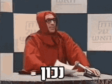 a man in a red robe is sitting at a desk with a pen and papers .