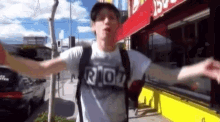 a man wearing a riot t-shirt is standing on a sidewalk with his arms outstretched .