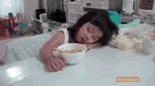 a little girl is sleeping at a table with a bowl of cereal in her hand .