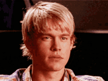 a young man with blonde hair and a plaid shirt looks at the camera