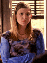 a woman in a blue tie dye shirt stands in front of a window with her arms crossed