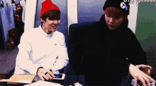 a man wearing a black hat with the letter x on it is sitting next to another man wearing a red hat