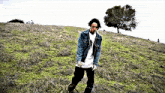 a man in a denim jacket is standing in a grassy field with a tree in the background
