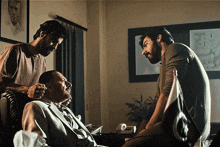 a man in a suit sits in a chair with two men standing around him