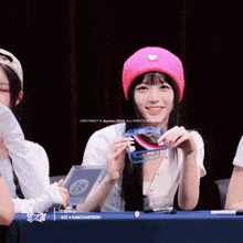 a girl wearing a pink hat with a heart on it is holding a phone