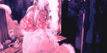 a woman with pink hair and a feathered headpiece looks at herself in a mirror