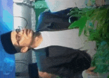 a man wearing glasses and a black hat is standing next to a plant ..