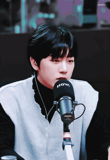 a young man wearing a black turtleneck and a white vest stands in front of a now microphone