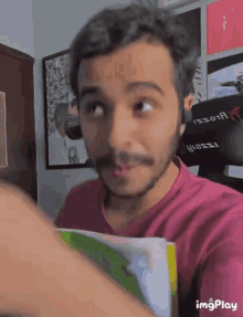 a man with a beard wearing a pink shirt is holding a book and making a face .