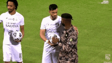a man in a military uniform holds a soccer ball in front of two men in saudia jerseys