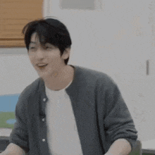 a young man wearing a gray cardigan and a white shirt is sitting at a table and smiling .