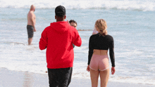 a man in a red hoodie takes a picture of a woman walking on the beach
