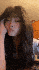 a woman with long dark hair and bangs is sitting on a bed with her hand on her face .
